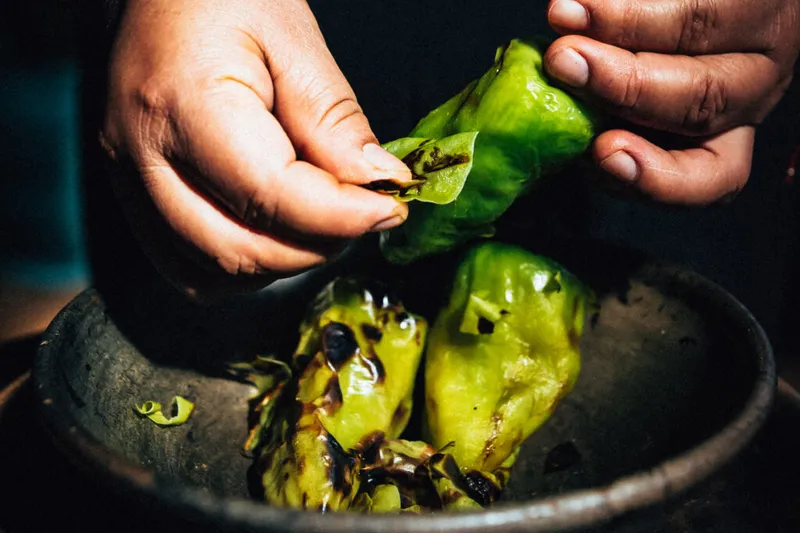 Hands peeling fire-blistered green chiles de agua over a dark clay molcajete at Madre! Oaxacan Restaurant and Mezcaleria