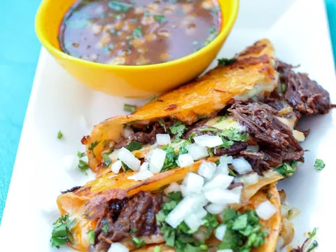 Three crispy quesabirria tacos filled with shredded beef, onion and cilantro on a white plate at Rev Mex, served with a yellow bowl of consommé for dipping