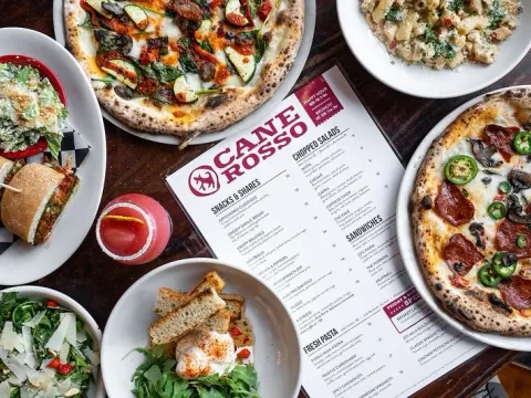 Overhead spread of wood-fired pizzas, salads, sandwiches and a cocktail around a Cane Rosso menu