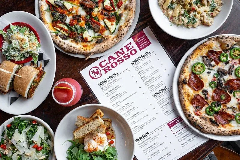 Overhead spread of wood-fired pizzas, salads, sandwiches and a cocktail around a Cane Rosso menu
