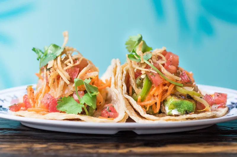 Two street-style tacos topped with pico de gallo, shredded carrot and crispy potato on a white plate at Broken English Taco Pub