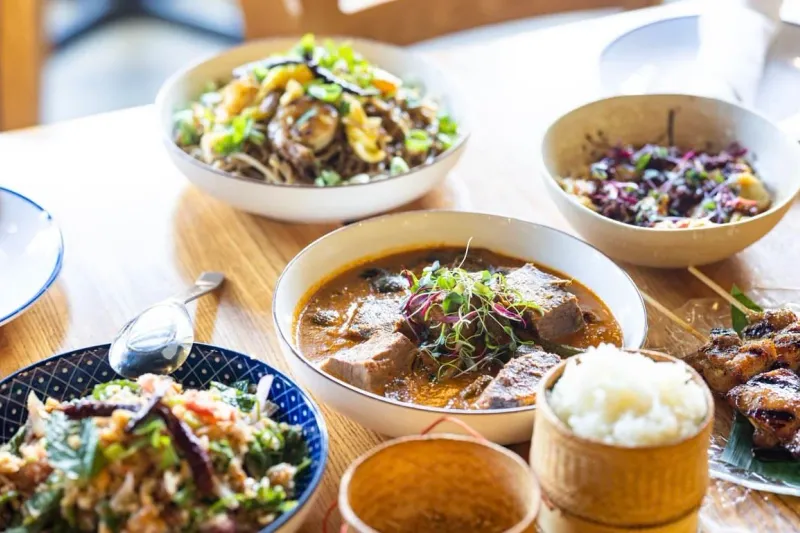 A spread of Lao dishes at Thip Khao including a curry, a minced-herb salad, stir-fried noodles, grilled skewers and woven bamboo baskets of sticky rice on a wooden table