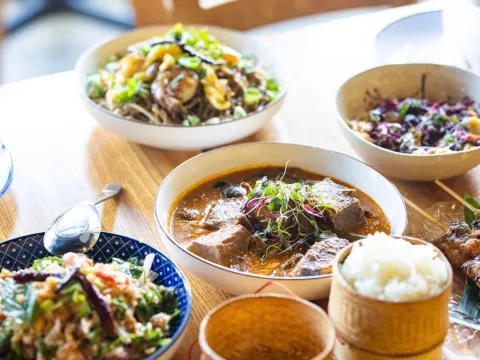 A spread of Lao dishes at Thip Khao including a curry, a minced-herb salad, stir-fried noodles, grilled skewers and woven bamboo baskets of sticky rice on a wooden table
