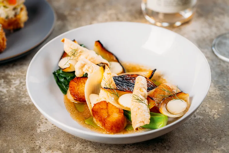 Seared scallops with mushrooms, greens, and broth in a white bowl at The Antler Room in Kansas City.