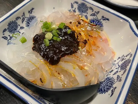 A blue-and-white porcelain dish of chilled translucent Sichuan noodles topped with chili sauce, dark bean sauce and scallions, with a Mala Restaurant chopstick sleeve behind it