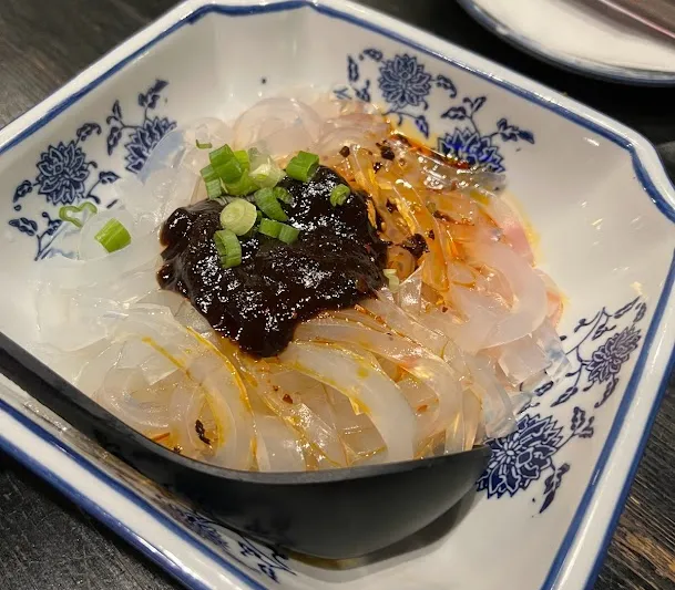 A blue-and-white porcelain dish of chilled translucent Sichuan noodles topped with chili sauce, dark bean sauce and scallions, with a Mala Restaurant chopstick sleeve behind it