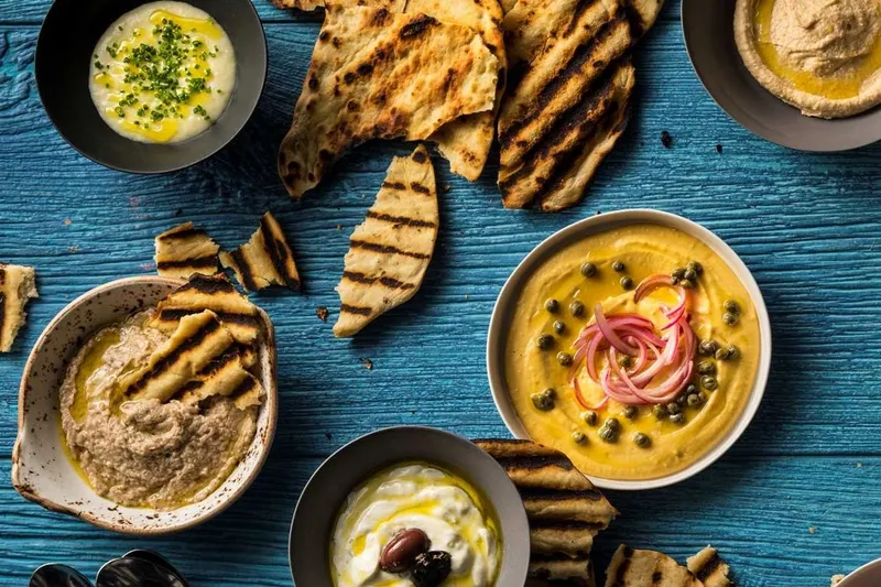 Overhead Greek meze spread at Kyma in Atlanta with bowls of whipped dips and spreads topped with capers and pickled onion, alongside charred grilled pita