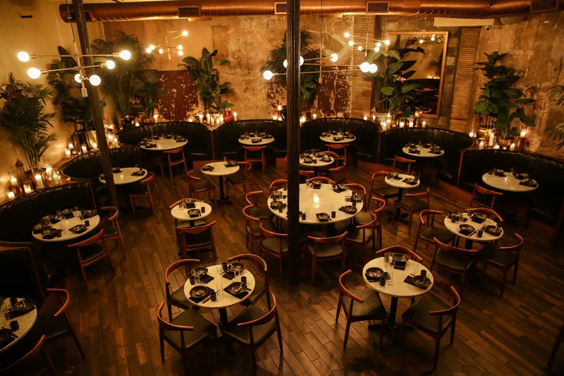 Candlelit dining room at Chinese Tuxedo with marble-topped tables, green leather banquettes, hanging bulb chandeliers and exposed brick walls