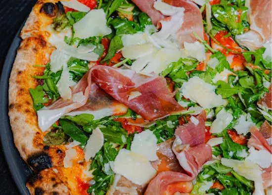 Close-up of Pizzana's Margherita al Crudo pizza topped with prosciutto di parma, arugula and shaved parmigiano on a dark pan