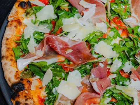 Close-up of Pizzana's Margherita al Crudo pizza topped with prosciutto di parma, arugula and shaved parmigiano on a dark pan