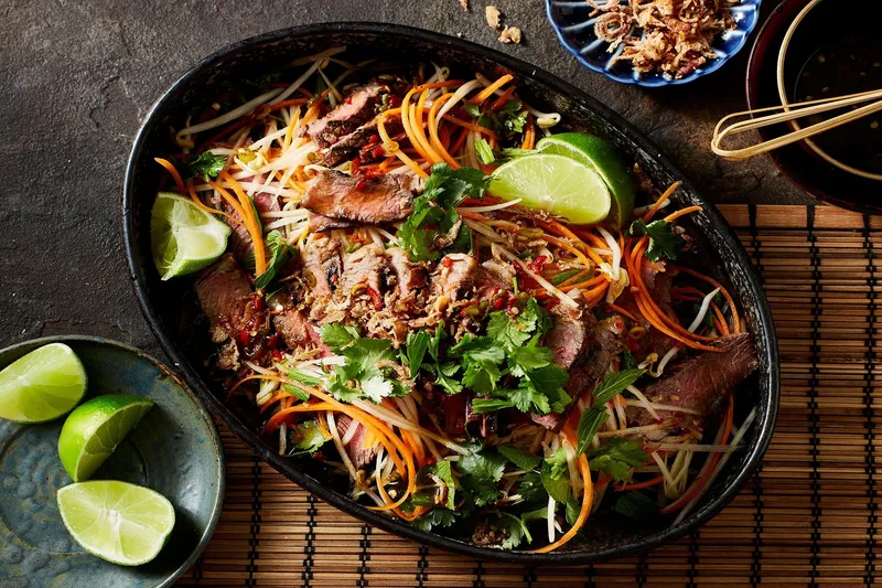 Thai grilled beef salad with shredded green papaya, carrots, herbs, chilies and lime wedges served in a dark oval dish at Bangrak Market in Seattle