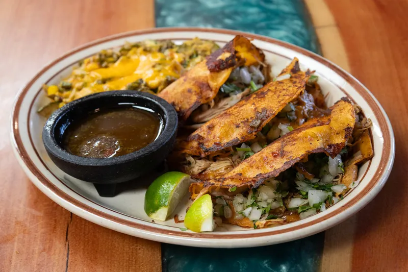 A plate of Sadie's of New Mexico green chile chicken birria tacos with melted cheese, onion and cilantro, served with a cup of consommé, calabacitas and lime wedges