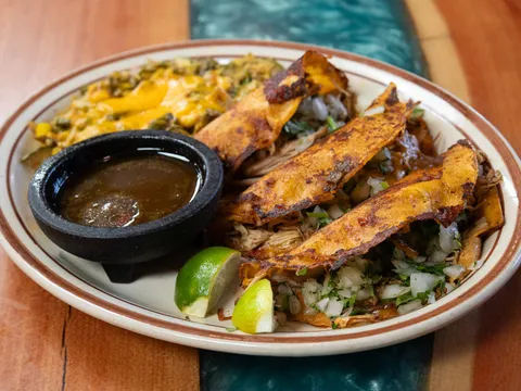 A plate of Sadie's of New Mexico green chile chicken birria tacos with melted cheese, onion and cilantro, served with a cup of consommé, calabacitas and lime wedges