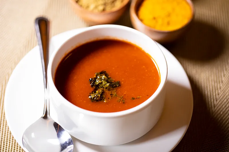 Bowl of tomato basil shorba garnished with herbs, with spice bowls behind, at The Yellow Chilli, Plano