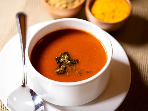 Bowl of tomato basil shorba garnished with herbs, with spice bowls behind, at The Yellow Chilli, Plano