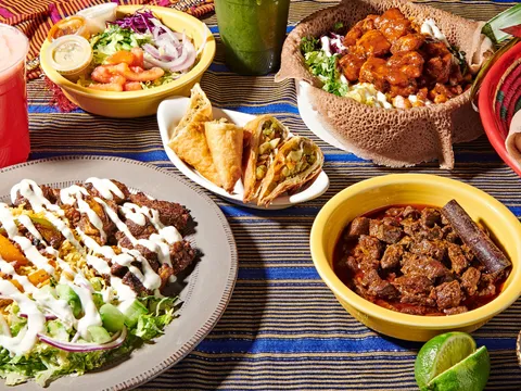 A spread of Somali dishes at Hoyo's Kitchen: a suqaar plate over yellow rice with salad, fried sambusas, injera with saucy chicken, a bowl of beef stew and a red juice on a striped tablecloth