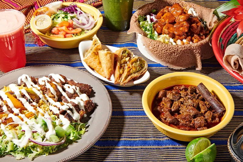 A spread of Somali dishes at Hoyo's Kitchen: a suqaar plate over yellow rice with salad, fried sambusas, injera with saucy chicken, a bowl of beef stew and a red juice on a striped tablecloth
