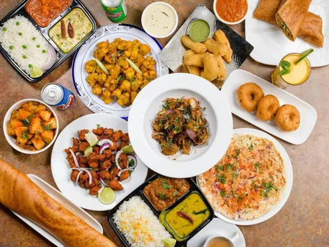 Overhead spread of South Indian dishes at NALA'S: a paper-thin dosa, idli and vada, thali trays with rice and curries, chicken 65, samosas, medu vada, mango lassi and a cup of masala chai
