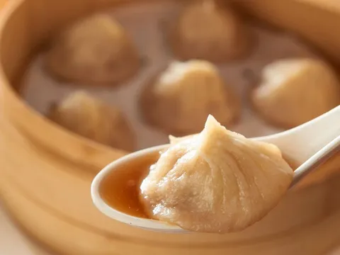 Royal China soup dumpling on a white spoon in front of a bamboo steamer.