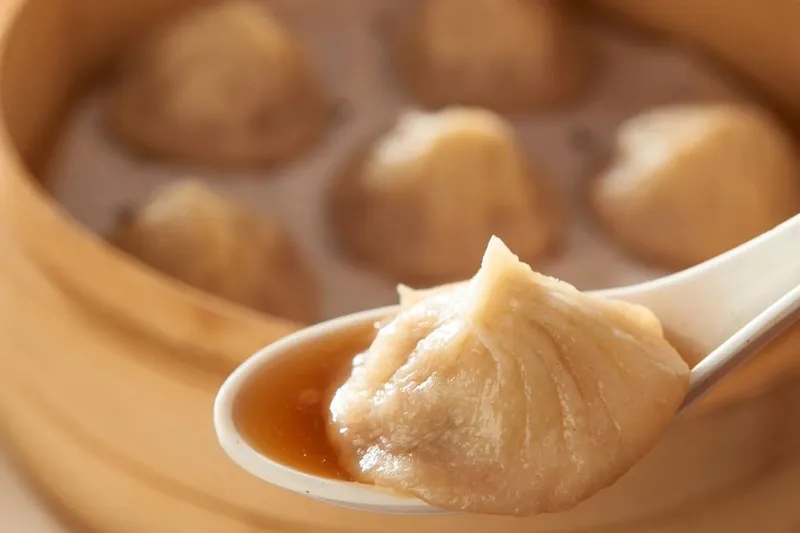 Royal China soup dumpling on a white spoon in front of a bamboo steamer.