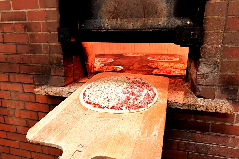 A raw apizza on a wooden peel in front of Modern Apizza's brick coal-fired oven with pizzas baking inside