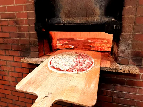 A raw apizza on a wooden peel in front of Modern Apizza's brick coal-fired oven with pizzas baking inside