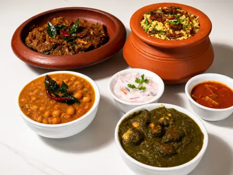 A spread of Mayura Indian Restaurant's Kerala dishes in clay pots and bowls: biryani, coconut roast, chana masala, raita, fish curry, and palak paneer