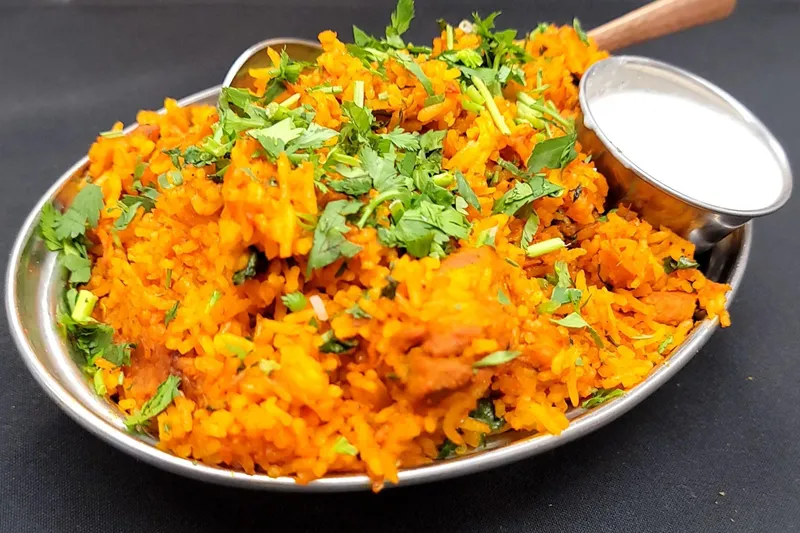 Chicken biryani topped with cilantro and served with a cup of raita in a steel dish at Little India Restaurant & Bar in Denver