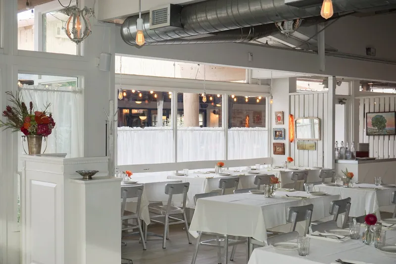 Bright, white-walled dining room at Tratto in Phoenix, with white-clothed tables, metal chairs, and a flower arrangement near the front windows