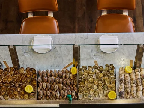 The raw-bar display at The Ordinary in Charleston: trays of oysters and clams on ice under glass at the marble counter, with leather bar stools above