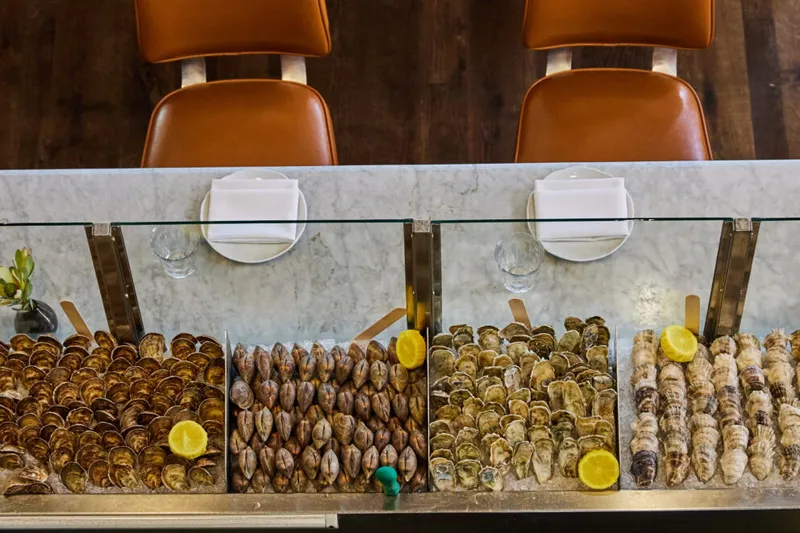 The raw-bar display at The Ordinary in Charleston: trays of oysters and clams on ice under glass at the marble counter, with leather bar stools above