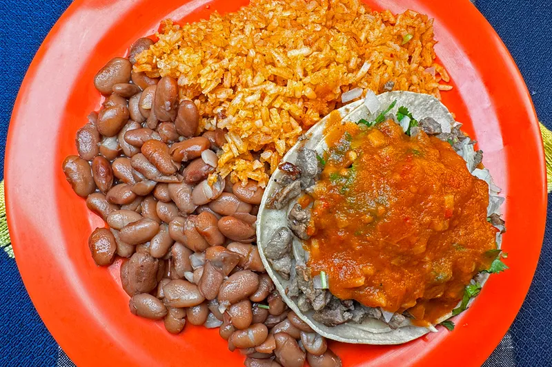 A carne asada taco topped with salsa alongside Mexican rice and pinto beans on an orange plate at Taquería Vallarta