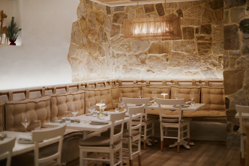 Dining room at Estiatorio Plaka in Baltimore's Greektown, with a rough stone wall, cream tufted banquettes, white wooden chairs and set tables under a woven pendant light