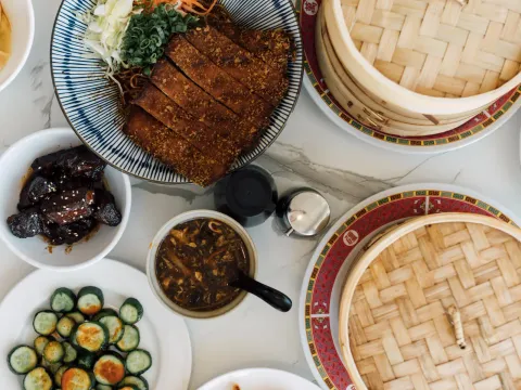 Overhead spread of China Max Dumpling House dishes on a marble table — crispy sliced pork chop over noodles, braised pork belly with sesame, chili cucumber salad, hot & sour soup, and bamboo steamers