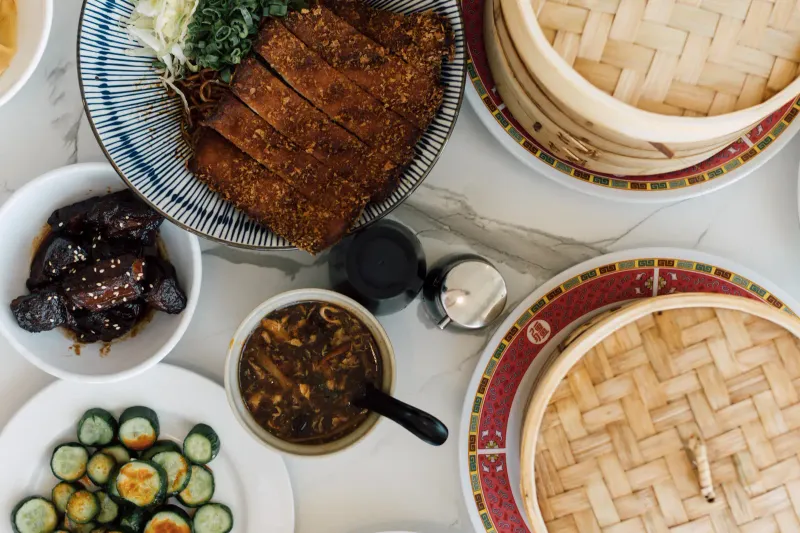 Overhead spread of China Max Dumpling House dishes on a marble table — crispy sliced pork chop over noodles, braised pork belly with sesame, chili cucumber salad, hot & sour soup, and bamboo steamers