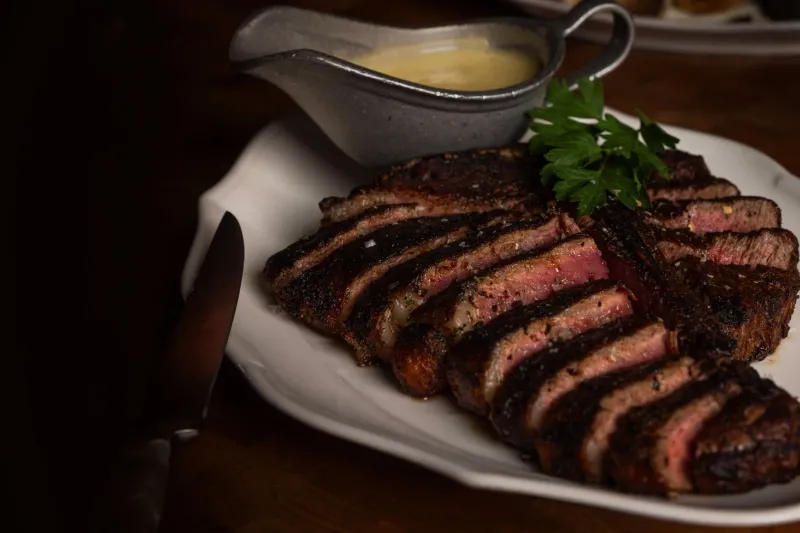 Sliced dry-aged steak on a white platter with a boat of béarnaise sauce and parsley at Bavette's Bar & Boeuf