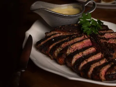 Sliced dry-aged steak on a white platter with a boat of béarnaise sauce and parsley at Bavette's Bar & Boeuf