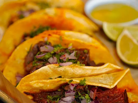 Close-up of Amor Y Tequila's tacos on soft corn tortillas topped with grilled meat, cilantro and red onion, with lime wedges and a cup of consommé