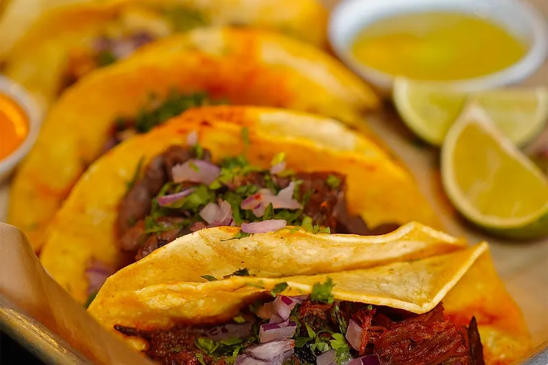 Close-up of Amor Y Tequila's tacos on soft corn tortillas topped with grilled meat, cilantro and red onion, with lime wedges and a cup of consommé