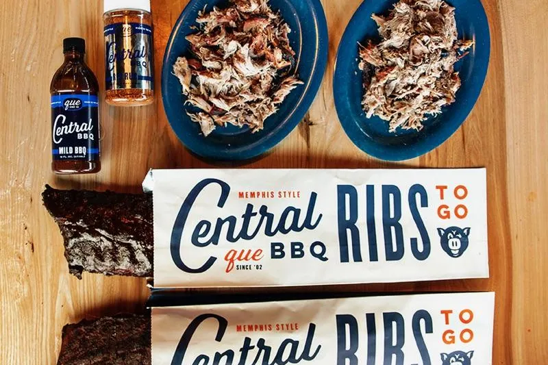 Central BBQ Memphis pulled pork on blue plates with smoked ribs, house BBQ sauce and dry rub, and branded Ribs To Go boxes