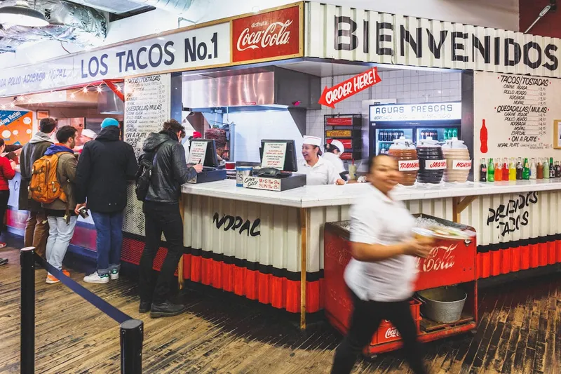 The Los Tacos No. 1 counter inside Chelsea Market, with the hand-painted 'Los Tacos No.1' and 'Bienvenidos' signs, aguas frescas jars, and customers ordering