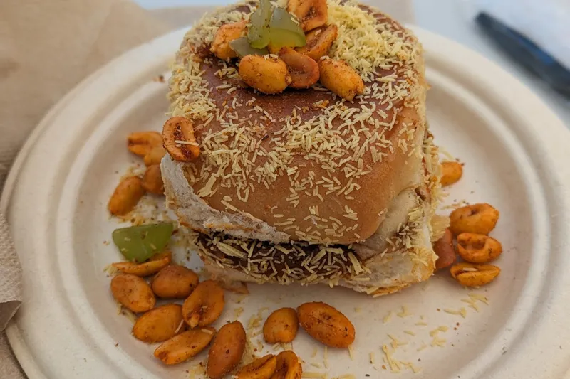 A pav (soft bun) street-food sandwich loaded with fine sev and spiced peanuts on a plate at Pav Bhaji and More, Sunnyvale