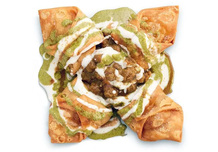Four crisp samosas arranged around chickpea chaat with green and white chutneys