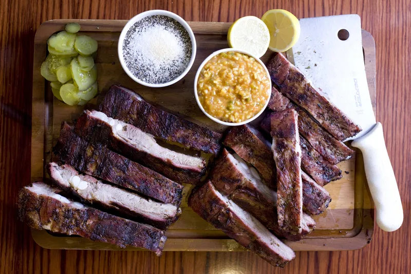 Overhead view of a Full Moon Bar-B-Que sliced pork rib slab on a wooden cutting board with dill pickles, dry rub, chow-chow relish, lemon halves and a butcher's cleaver