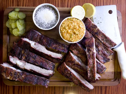 Overhead view of a Full Moon Bar-B-Que sliced pork rib slab on a wooden cutting board with dill pickles, dry rub, chow-chow relish, lemon halves and a butcher's cleaver
