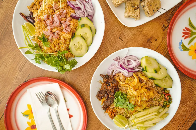 Overhead view of two Thai fried-rice dishes at Sri Siam Cafe with ham, cucumber, red onion, cilantro and lime, plus a plate of grilled tofu skewers on a wooden table