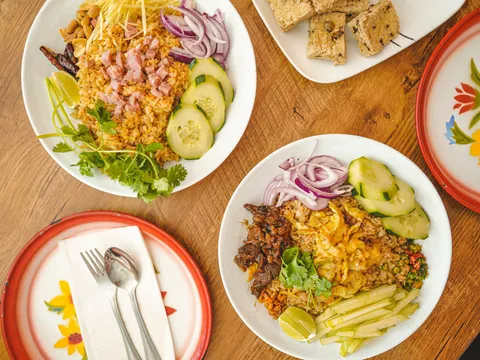 Overhead view of two Thai fried-rice dishes at Sri Siam Cafe with ham, cucumber, red onion, cilantro and lime, plus a plate of grilled tofu skewers on a wooden table