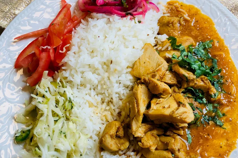 Chicken curry over basmati rice with cabbage slaw, sliced tomato and pickled red onions on a white plate at Klay Oven Kitchen