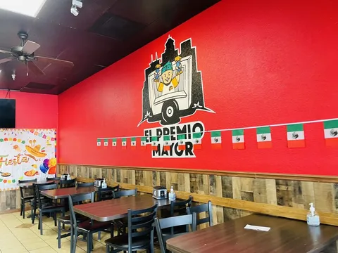 Dining room at El Premio Mayor in Fresno, with a hand-painted El Premio Mayor taco-truck mural on a red wall, wood-paneled wainscoting, wooden tables and chairs, and Mexican flag bunting