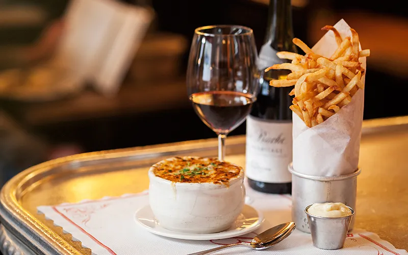 Balthazar's onion soup gratinée with a cone of pommes frites and a glass of red wine on a bistro tray
